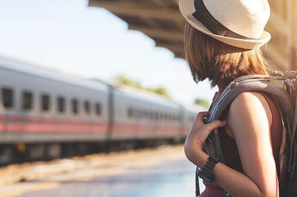 Frau mit Rucksack und Hut am Bahnsteig bei einfahrendem Zug