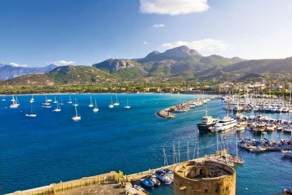 Malerischer Hafen von Calvi auf Korsika mit weißen Booten und blauem Meer vor mit Macchia bewachsenen Bergen